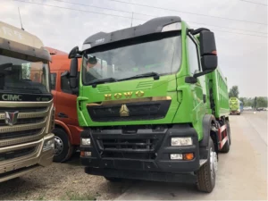 The image shows a green HOWO dump truck parked beside other trucks in an industrial yard