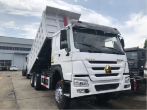 The image shows a white dump truck with its cargo bed lifted, parked outside a warehouse or factory