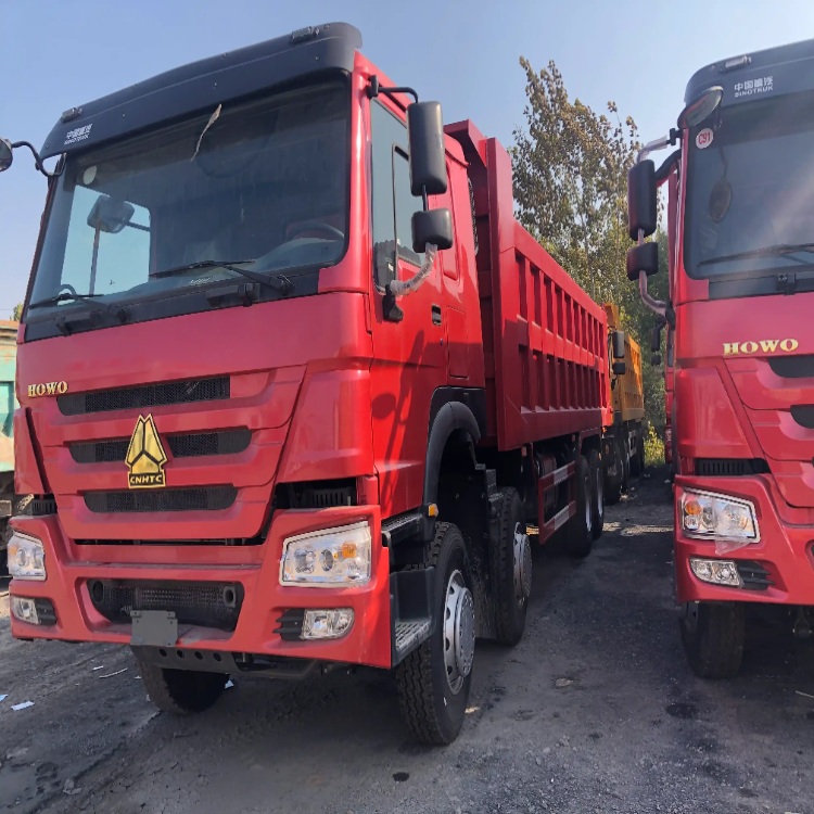 A red HOWO dump truck parked in an outdoor truck yard, viewed from thefront-left side, with another red truck beside it